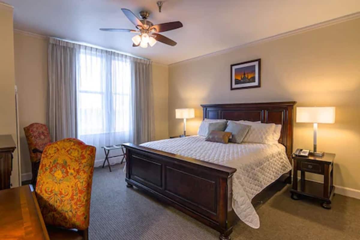 Une chambre d’hôtel avec un grand lit, deux tables de chevet, une chaise, un ventilateur de plafond et une fenêtre avec des rideaux.