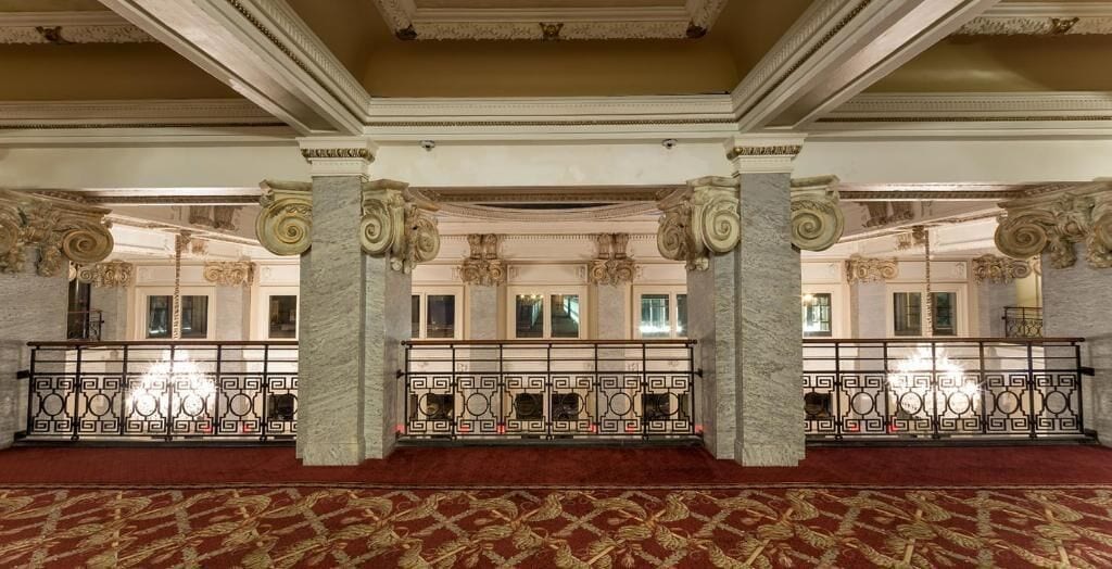A grand hall with ornate columns, a patterned carpet, and a balcony with a wrought iron railing.