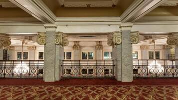 A grand hall with ornate columns, a patterned carpet, and a balcony with a wrought iron railing.