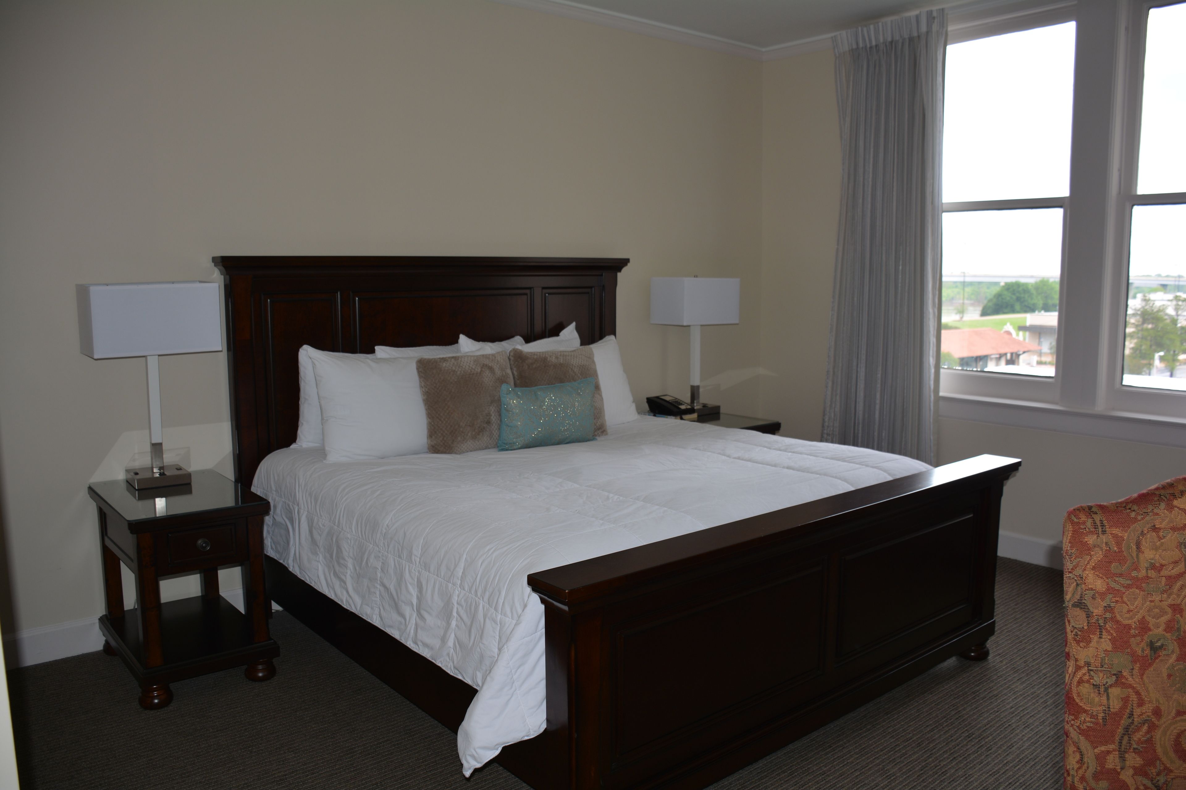 A hotel room with a large bed, two bedside tables with lamps, a window with a view, and a patterned armchair.