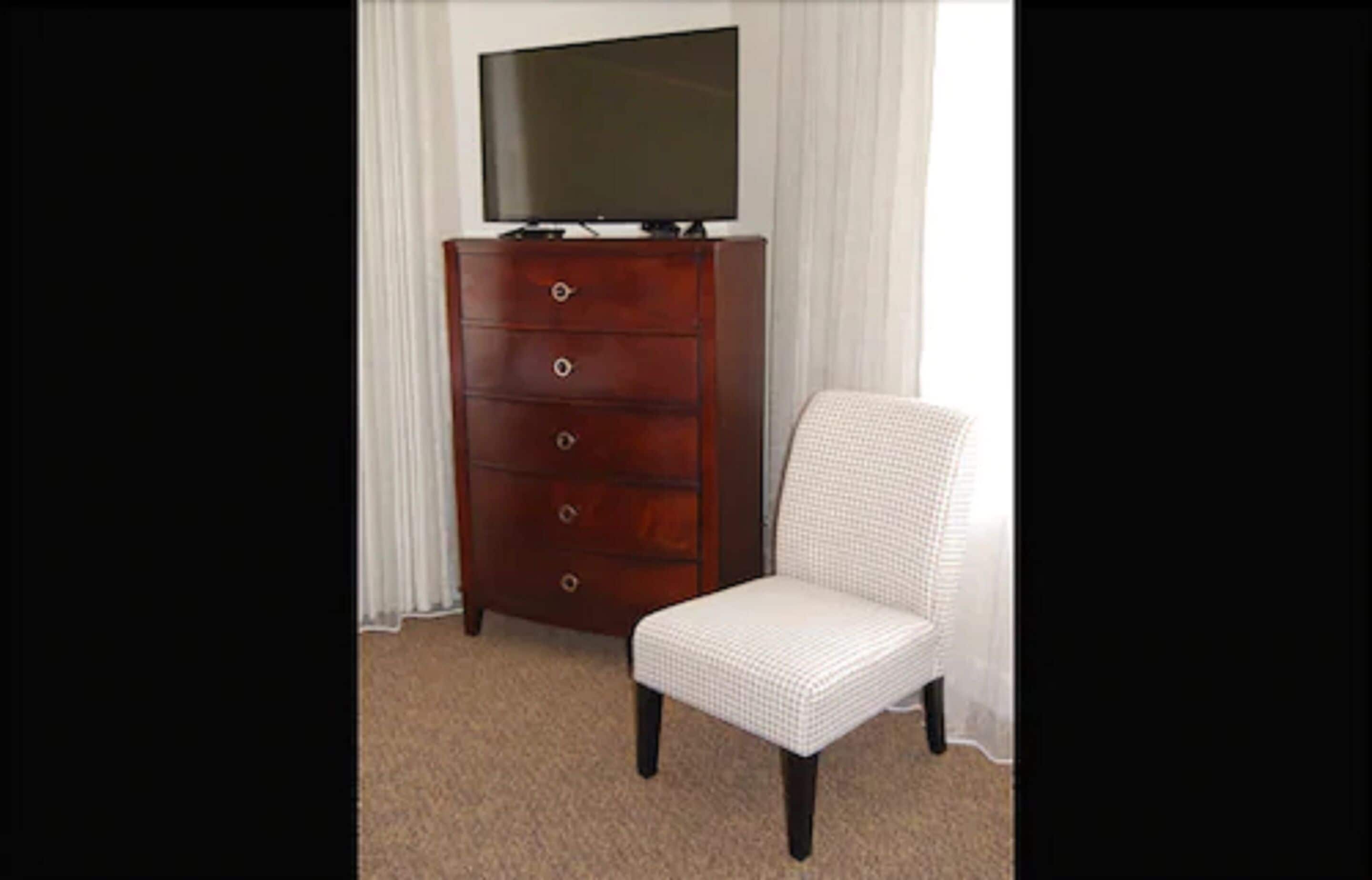 A wooden dresser with a television on top, a white chair, and a window with sheer curtains.