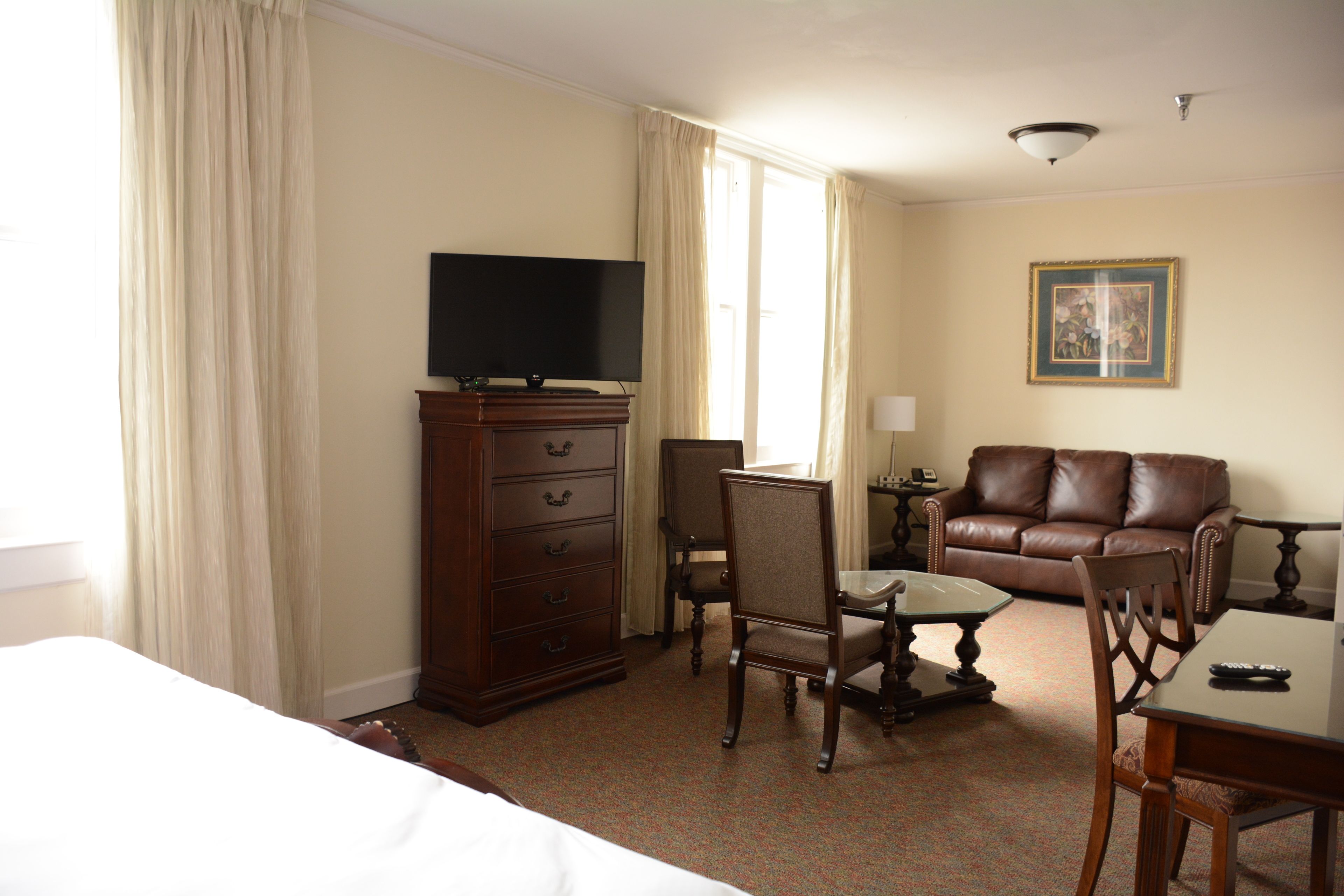 A hotel room with a bed, a television on a dresser, a brown leather sofa, a dining table with chairs, and a framed picture on the wall.