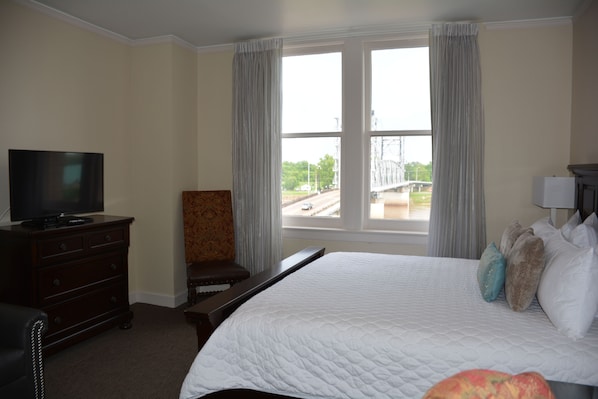 Une chambre d’hôtel avec un grand lit, un téléviseur posé sur une commode, une chaise et une fenêtre donnant sur un pont et une route.