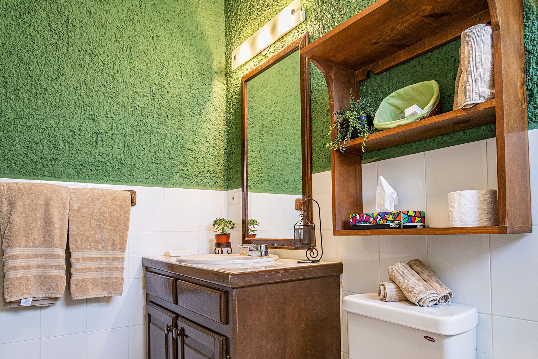 A bathroom with a green textured wall, a wooden vanity, a white toilet, and a shelf with towels and toiletries.
