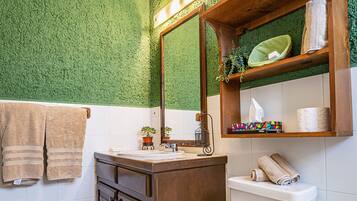 A bathroom with a green textured wall, a wooden vanity, a white toilet, and a shelf with towels and toiletries.