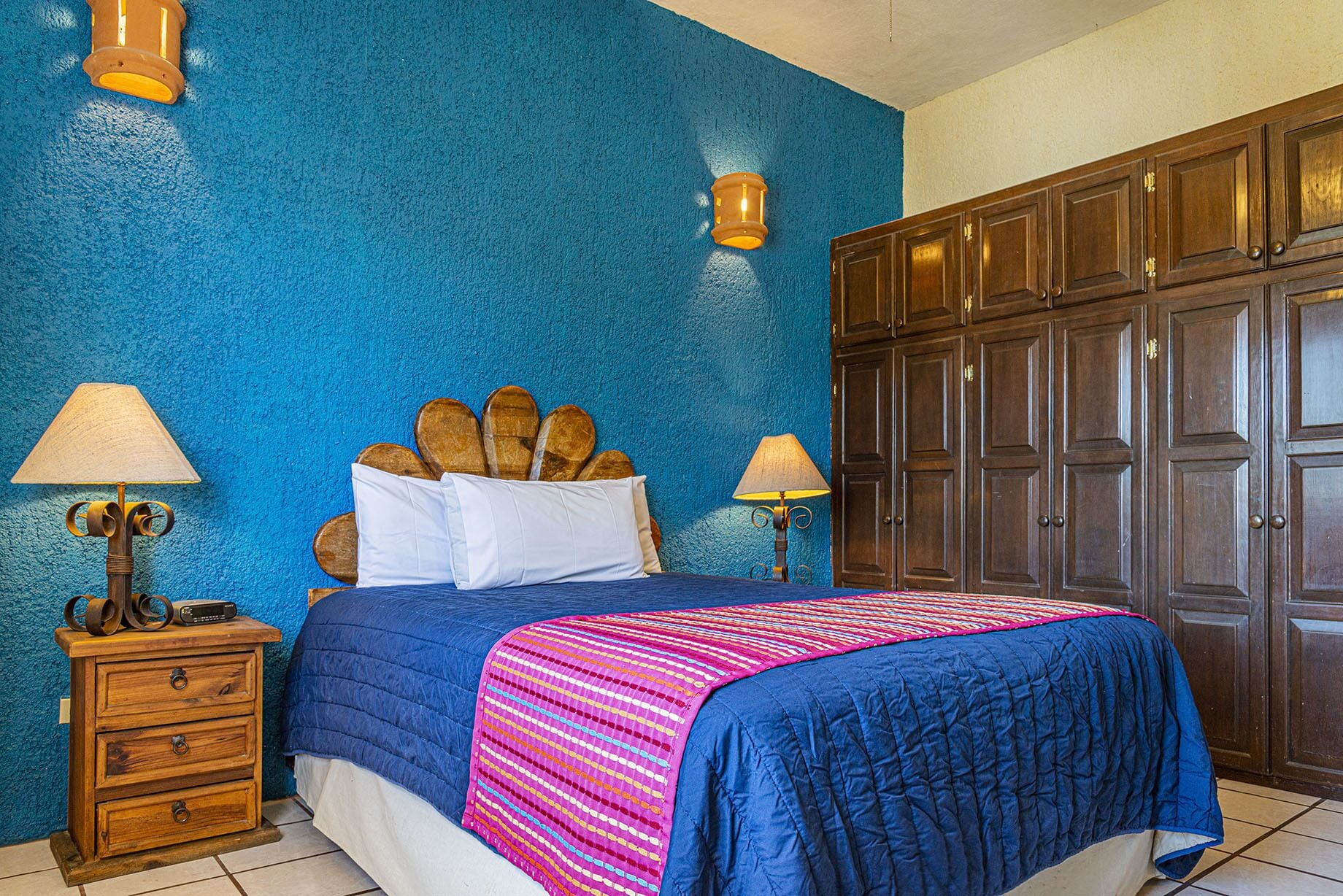 Une chambre à coucher avec un lit, deux tables de chevet avec des lampes, une armoire en bois et un mur bleu à texture.