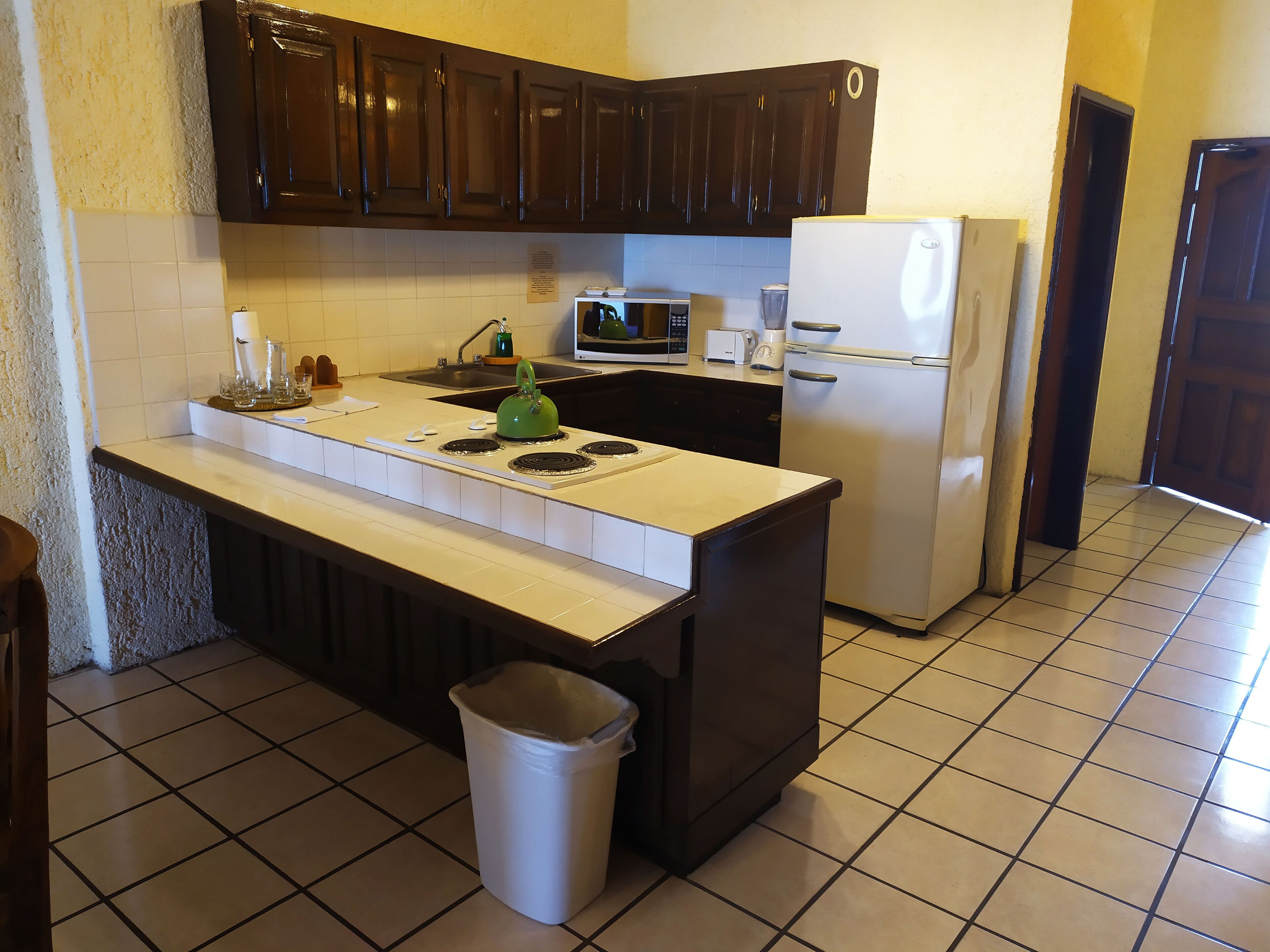 Una cocina con armarios oscuros, una encimera blanca, estufa, microondas, refrigerador y un cubo de basura.