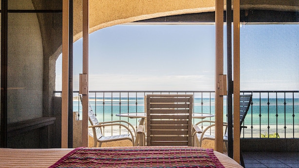 A balcony with a bed, two chairs, and a view of the beach and ocean.
