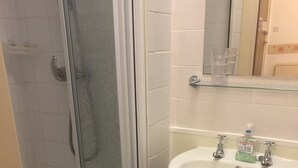 A bathroom with a glass shower enclosure, a white sink with two faucets, and a small trash can. - Brooklands Over 50's Only (Blackpool)