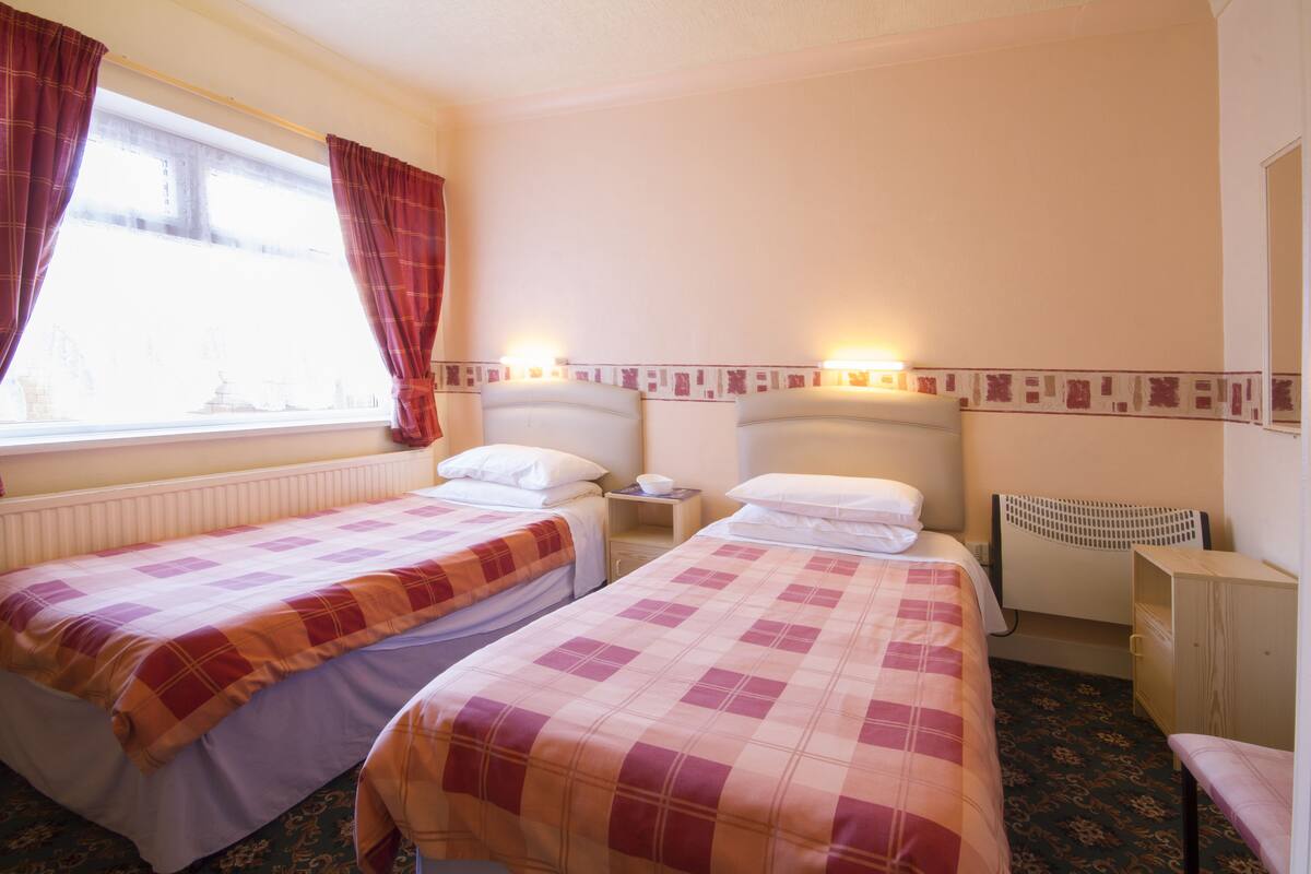 A hotel room with two beds, a window with curtains, a bedside table with a lamp, and a small cabinet.