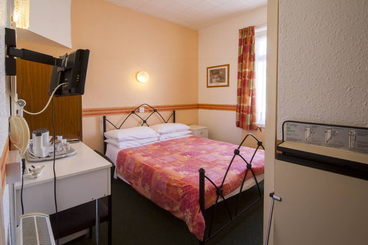 A hotel room with a bed, bedside table, lamp, and a small desk with a telephone and a coffee maker.
