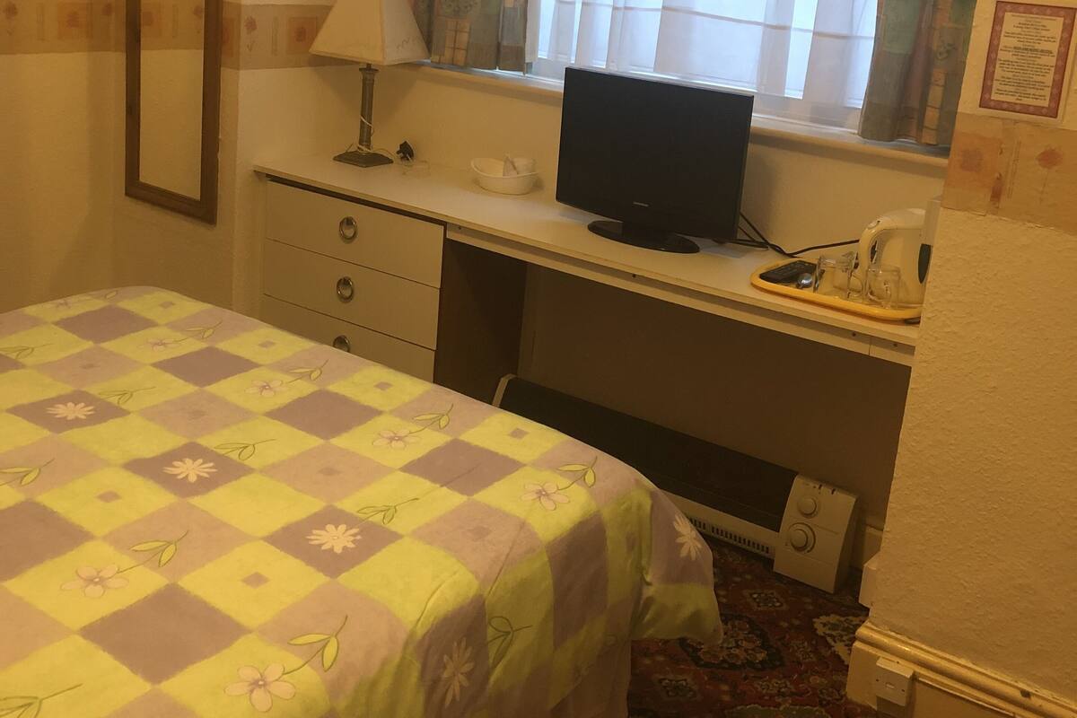 A bedroom with a bed, a desk with a computer, a window with curtains, a mirror, and a wall-mounted telephone.