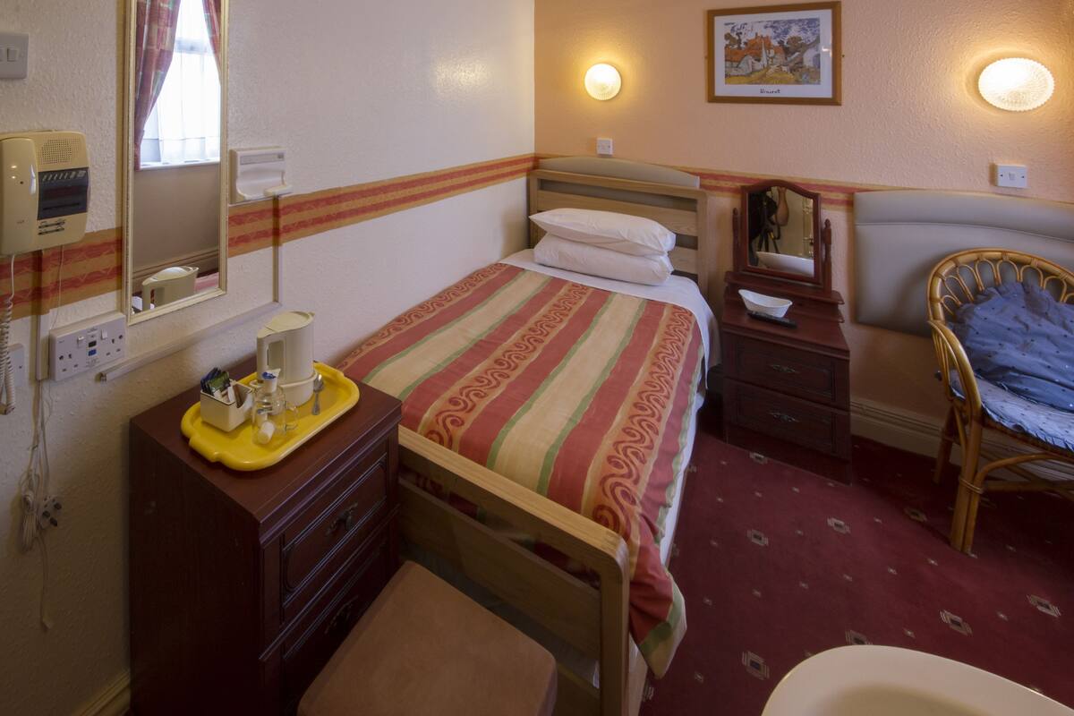 A hotel room with a bed, a wooden chair, a bedside table with a lamp, a mirror, and a wall-mounted phone.