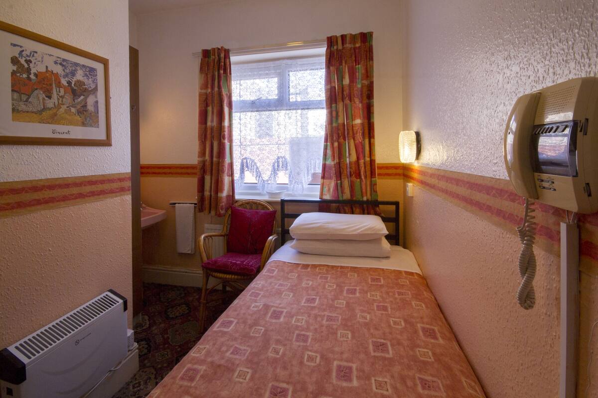 A hotel room with a bed, a chair, a desk, a telephone, and a window with curtains.