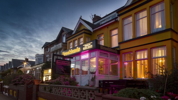A brightly lit hotel with a neon sign, a well-maintained garden, and a classic architectural style. - Brooklands Over 50's Only (Blackpool)