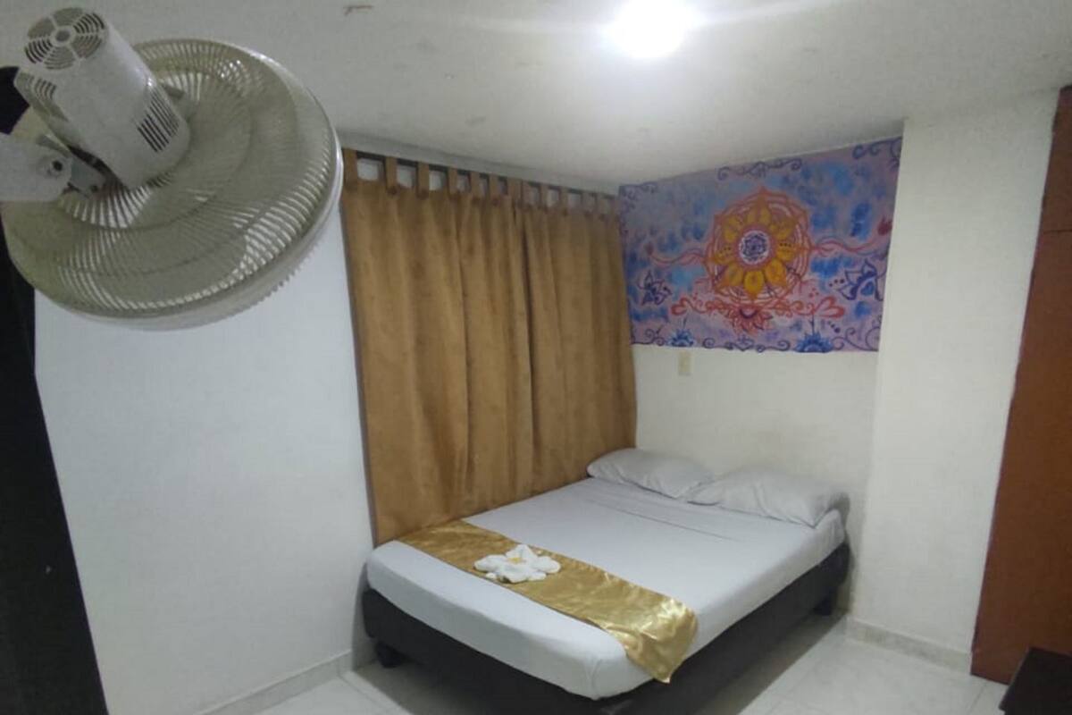 Une petite chambre propre, avec un lit simple, un ventilateur sur pied et une tapisserie colorée accrochée au mur.