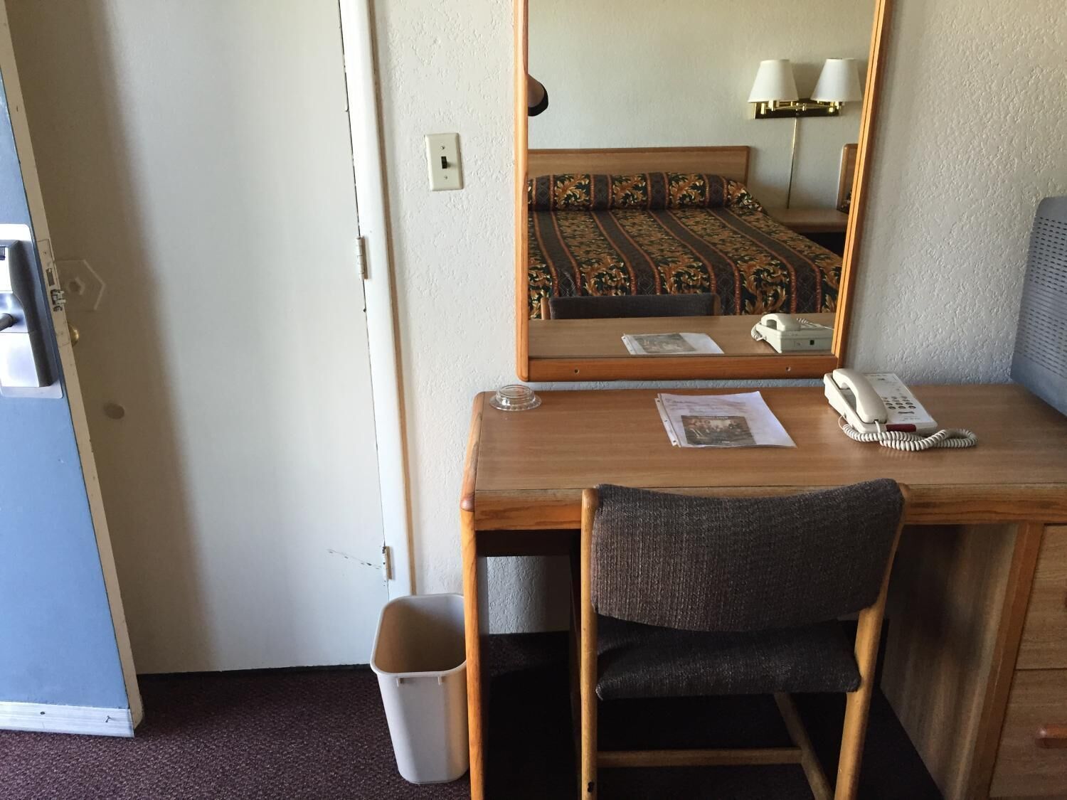 A hotel room with a wooden desk, a chair, a telephone, a mirror, and a small trash bin.