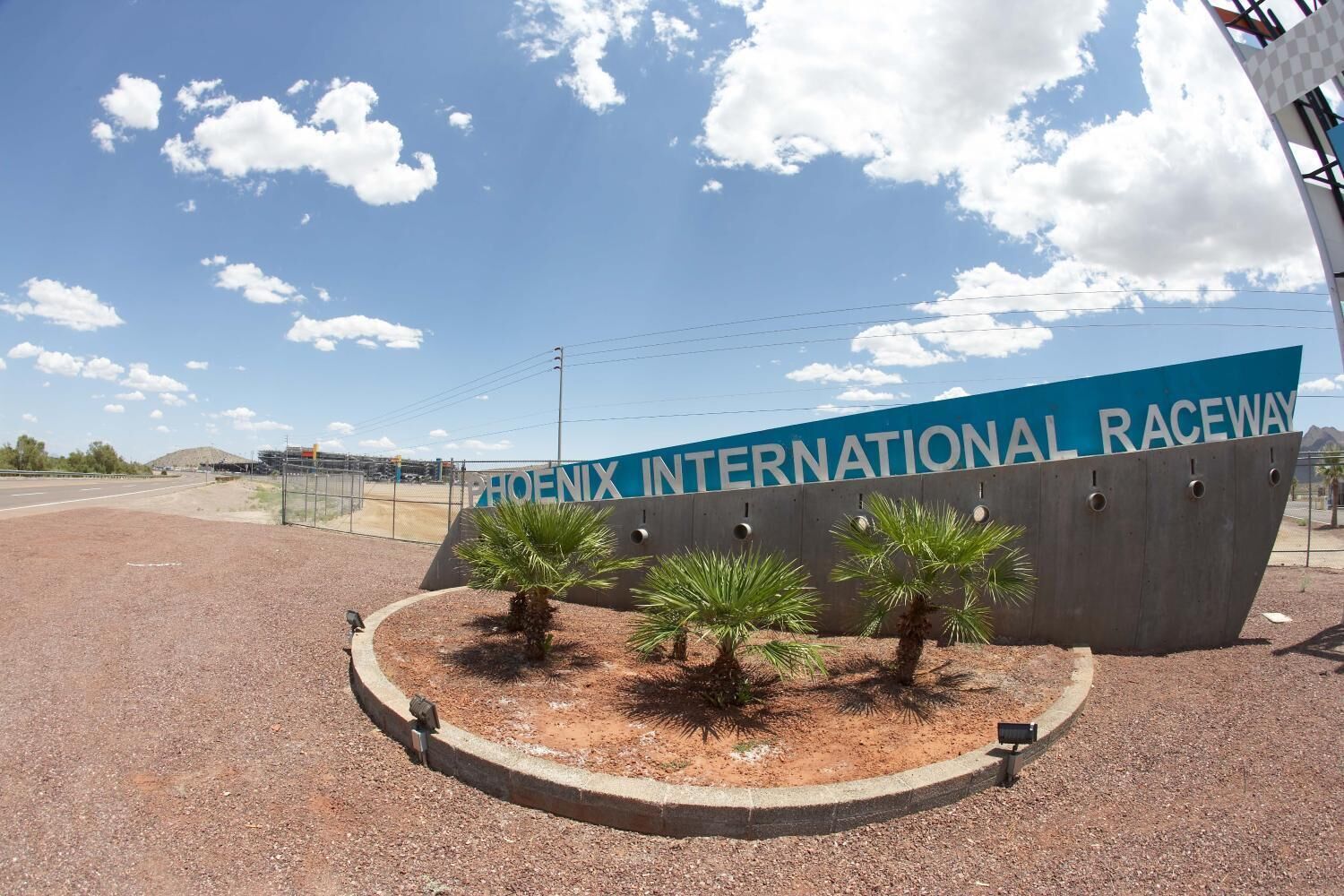 Une enseigne indiquant l’emplacement du circuit Phoenix International Raceway, avec un ciel dégagé et une zone de gravier bordée de palmiers.