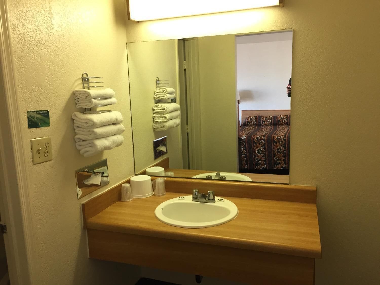 A hotel bathroom with a sink, mirror, towel rack, and a view of a bedroom with a patterned bedspread.