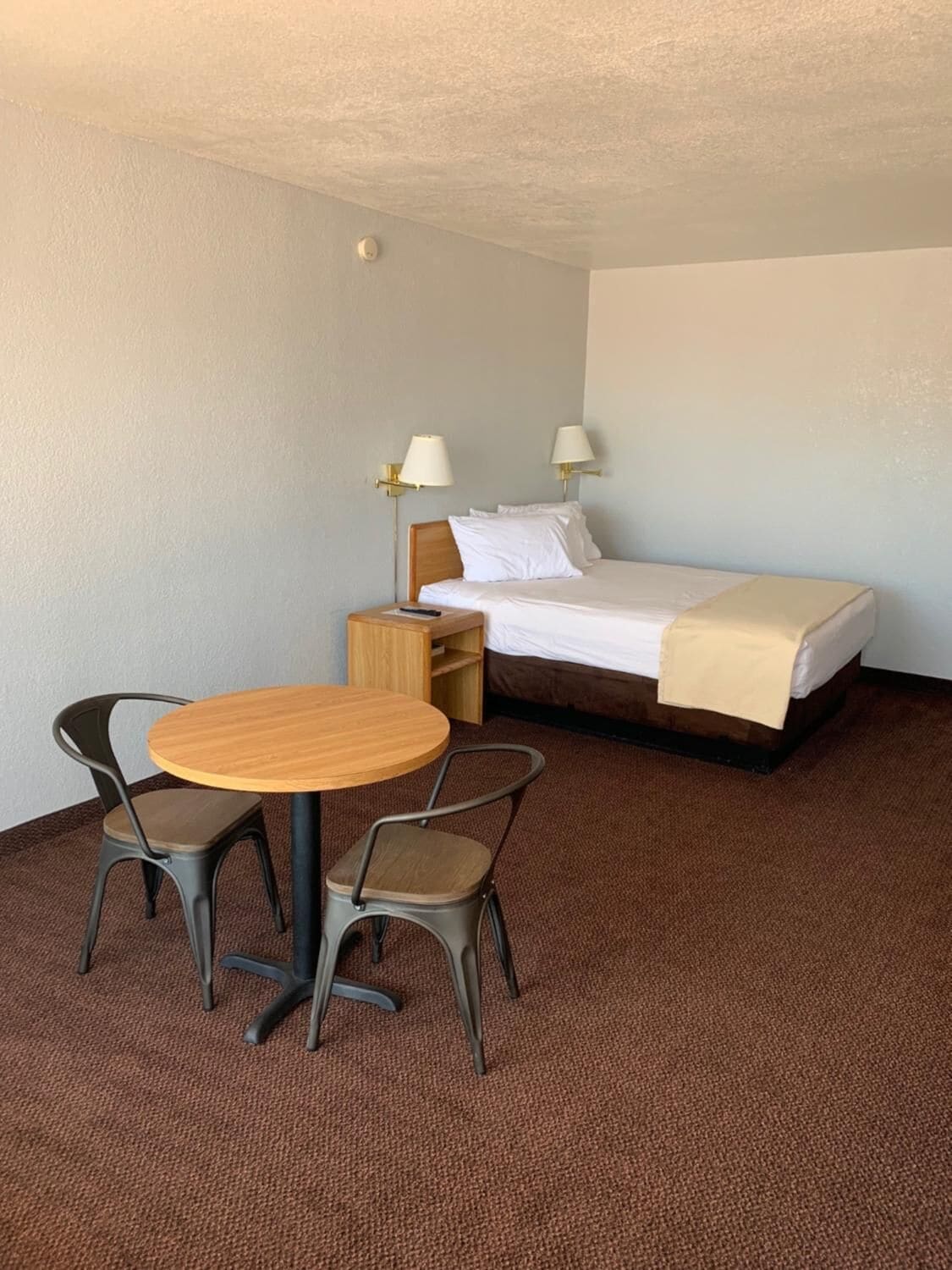 A hotel room with a single bed, two bedside lamps, a small wooden table with two chairs, and a carpeted floor.