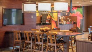 A hotel dining area with a wooden table, chairs, a fruit bowl, and a TV mounted on the wall. - Fairfield Inn and Suites by Marriott Chillicothe (Chillicothe)