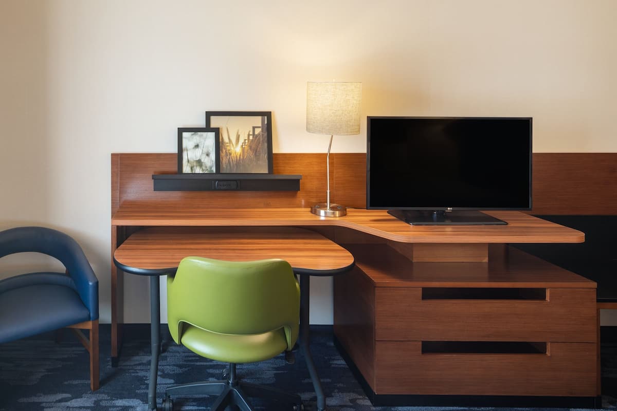 A hotel room with a wooden desk, a television, a lamp, a green chair, and a blue chair.