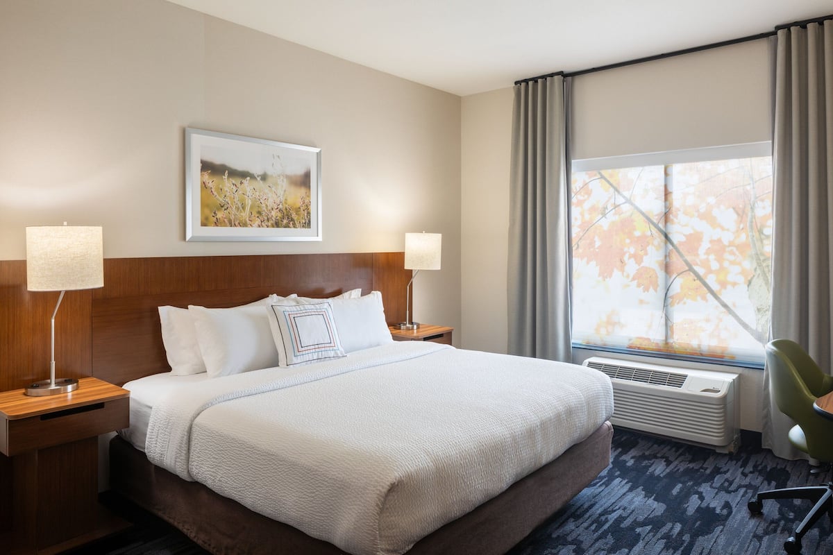 A hotel room with a large bed, two bedside tables with lamps, a window with curtains, a radiator, and a chair.