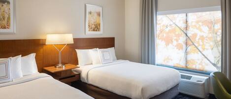 A hotel room with two beds, a nightstand, a lamp, a window with curtains, and a radiator.