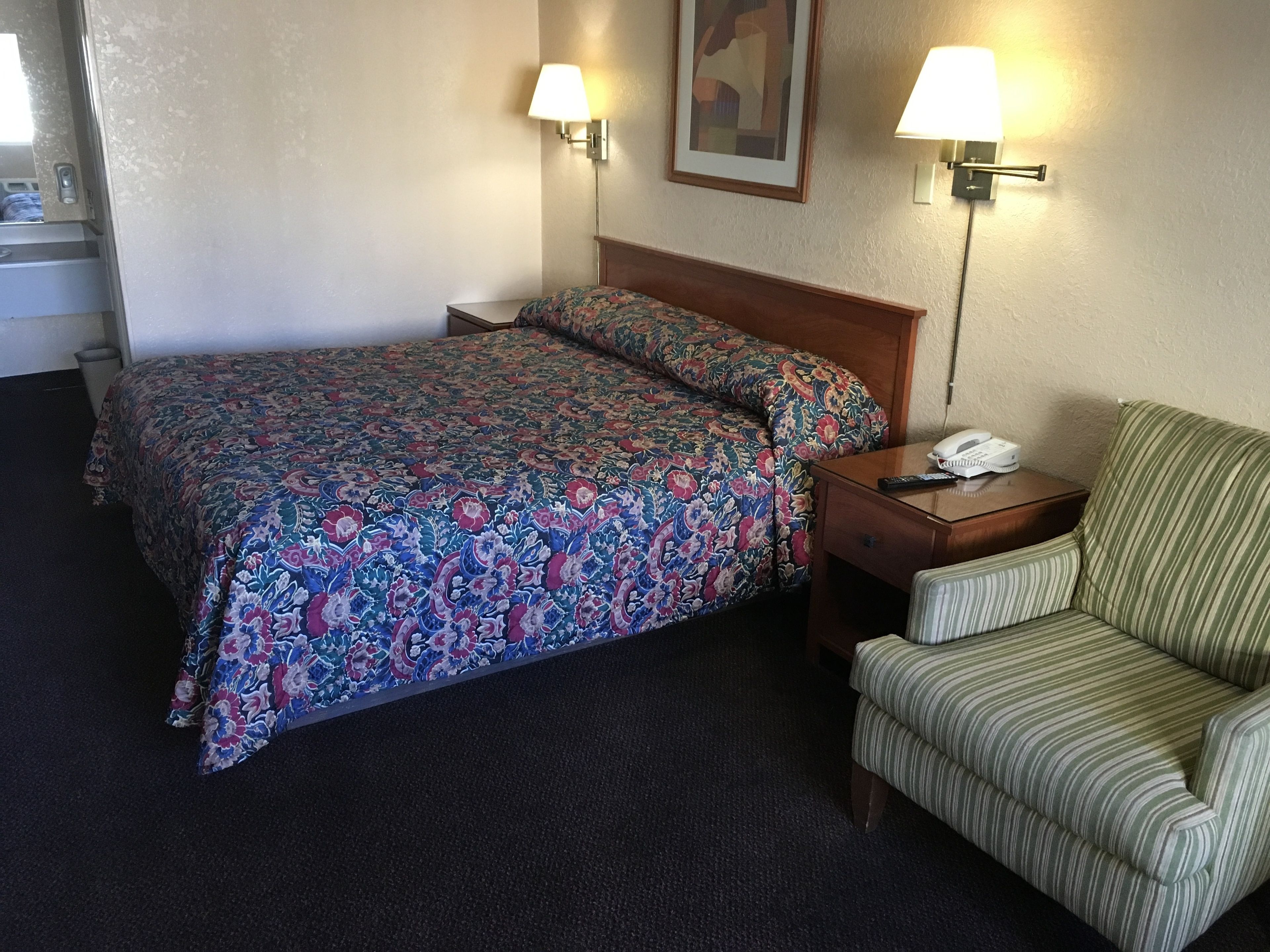 A hotel room with a bed, a chair, a nightstand with a phone, and two wall lamps.