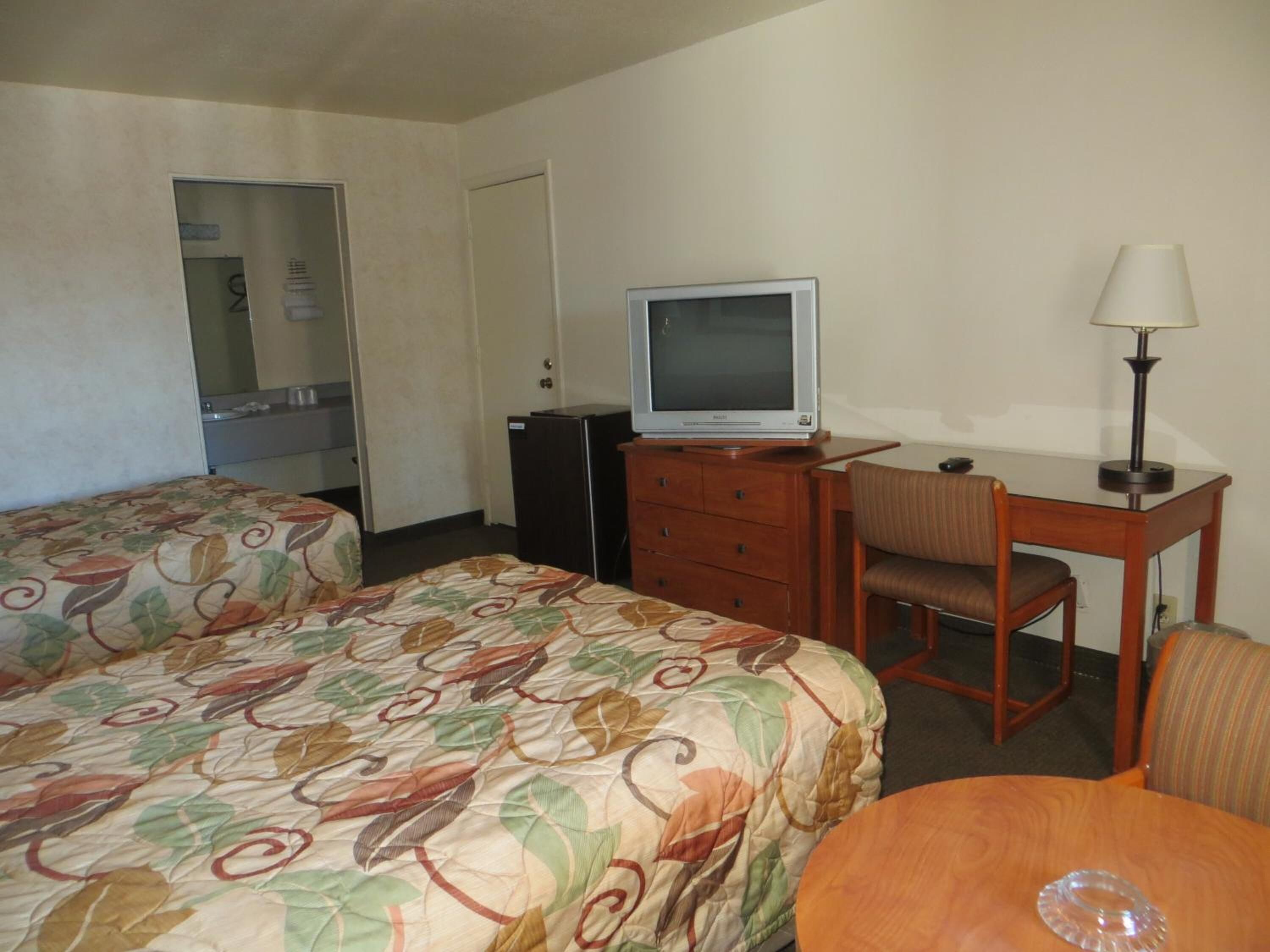 A hotel room with two beds, a desk with a chair, a television, a refrigerator, and a lamp.
