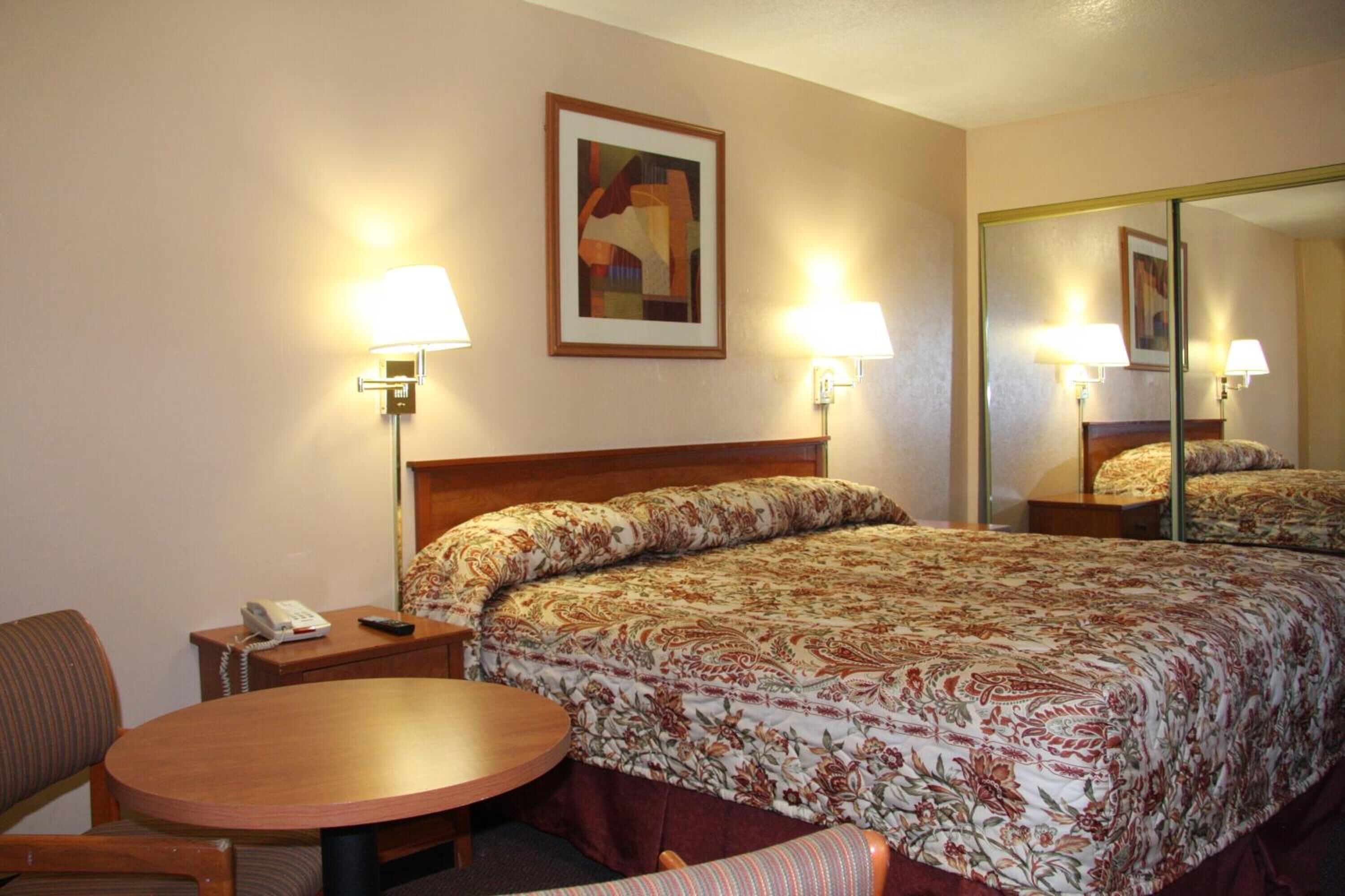 A hotel room with a bed, a round table, two lamps, a phone, and a painting on the wall.