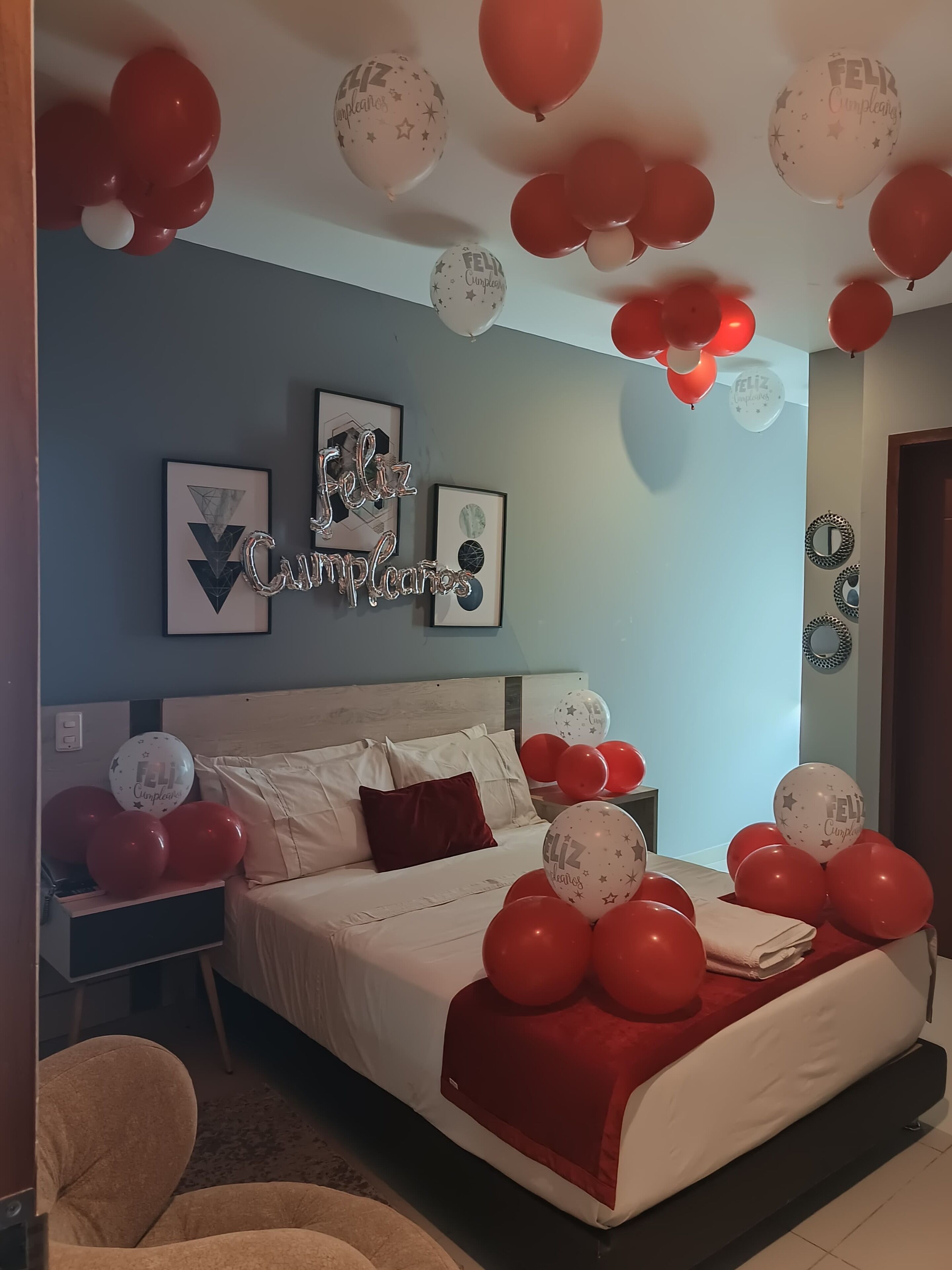 A bedroom with a bed, pillows, and a bedside table. There are balloons with the text "Feliz Cumpleaños" on the ceiling.