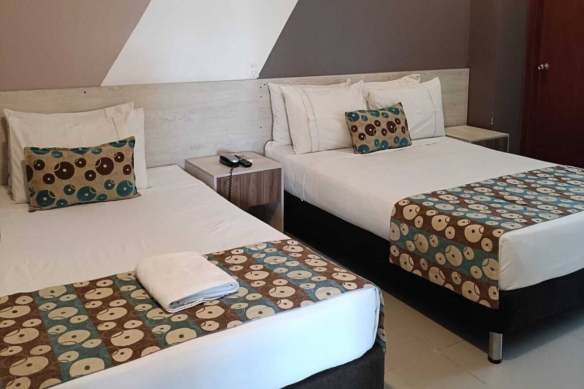 A hotel room with two beds, each with a patterned bedspread, a nightstand with a phone, and a door to the right.