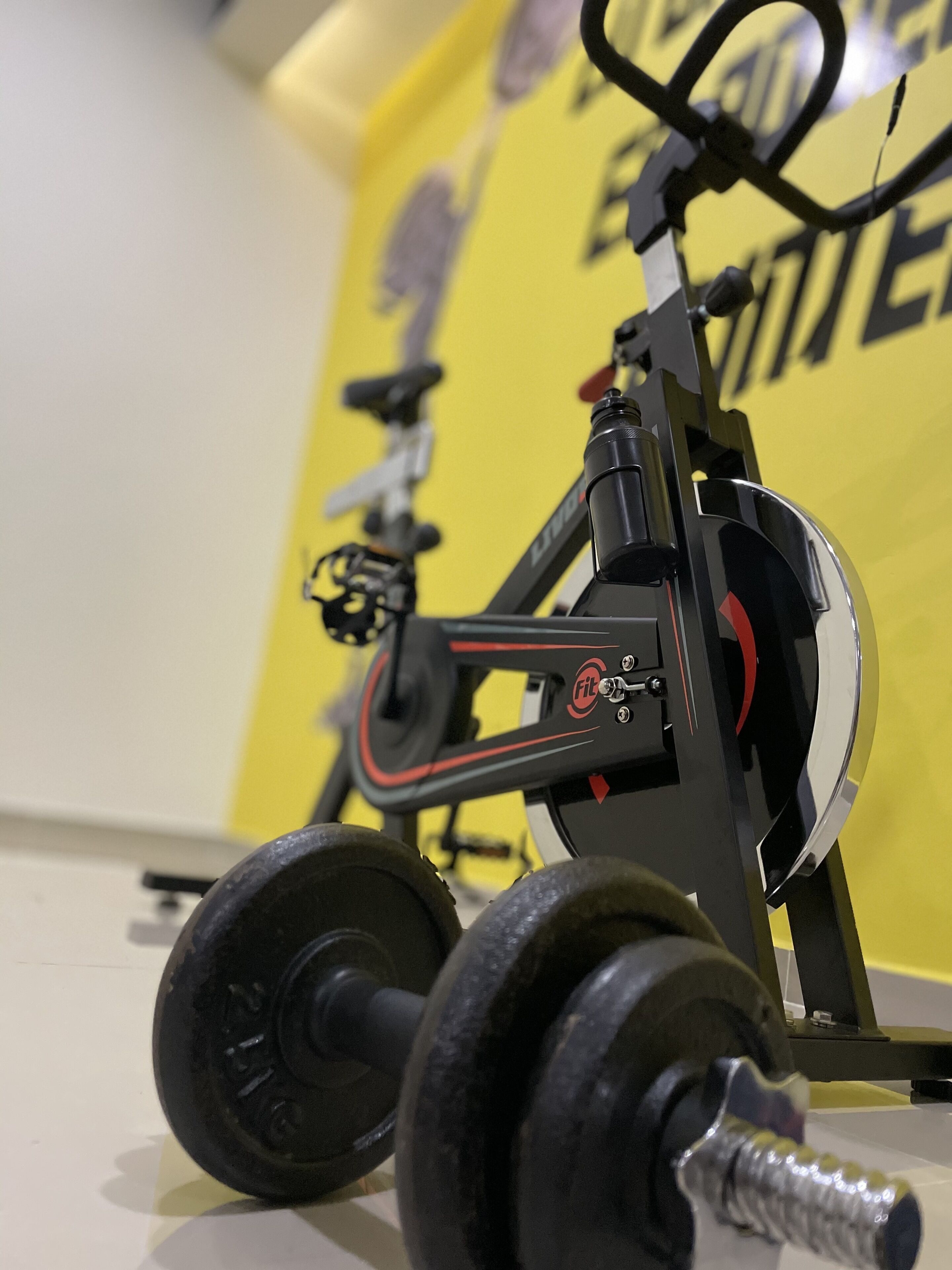 Uma bicicleta ergométrica fixa com estrutura preta e detalhes vermelhos, posicionada em frente a uma parede amarela, com uma visão parcial de uma bicicleta.