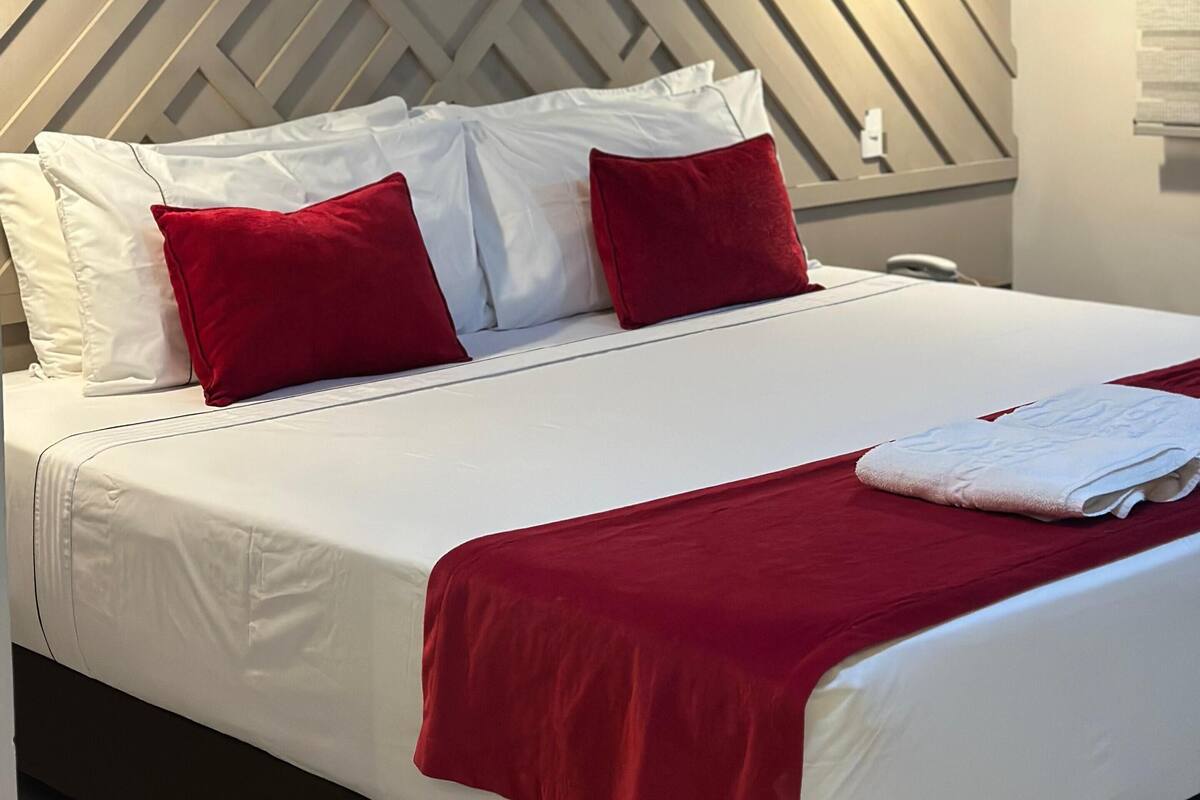 A modern hotel room with a geometric-patterned headboard, a neatly made bed with white linens and red accents, and a single bedside lamp.