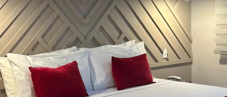 Una habitación de hotel moderna con cabecera de diseño geométrico, cama bien hecha con ropa blanca y detalles rojos, y una lámpara de noche.