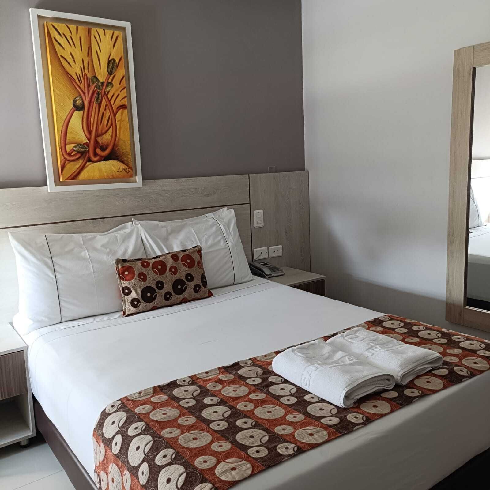 Un lit bien fait, avec des draps blancs, un plaid à motifs et un oreiller orné d’un motif circulaire. Un tableau est visible au mur, au-dessus du lit.