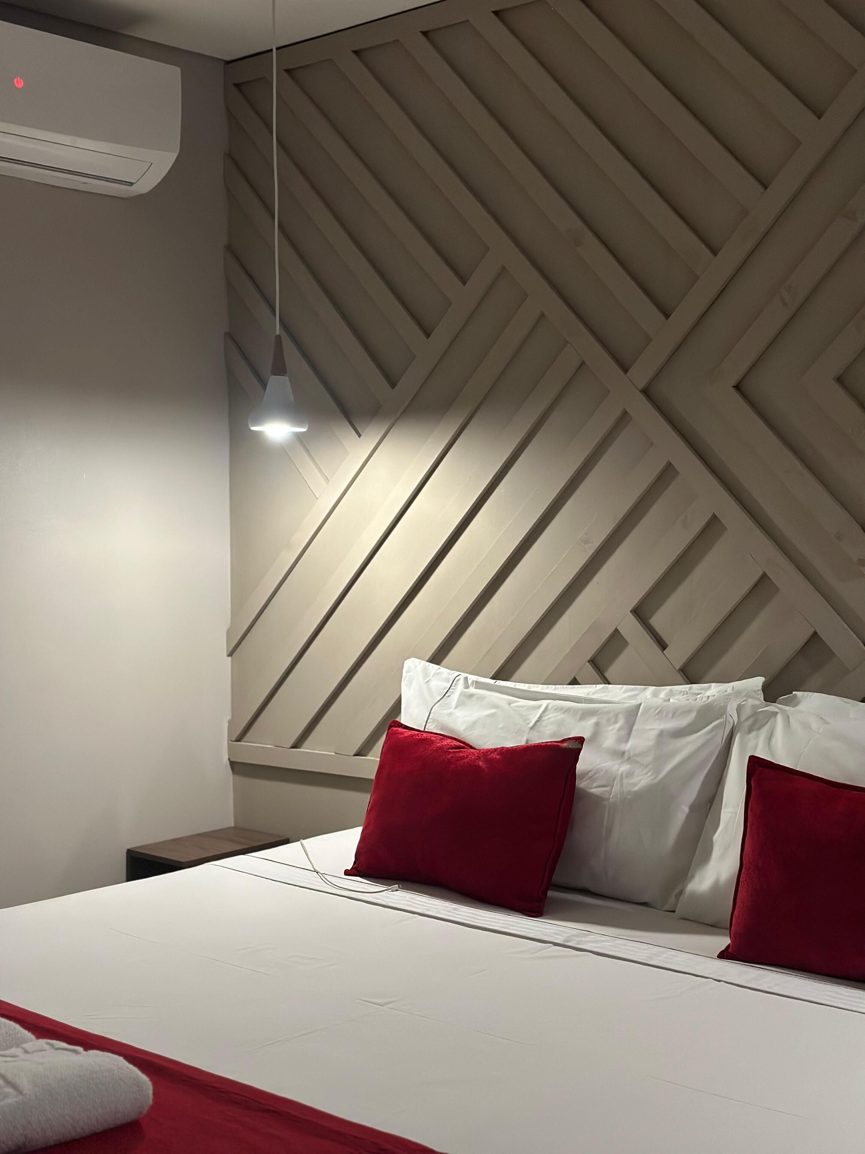 Ein modernes Schlafzimmer mit einem Bett, roten und weißen Kissen, einer Hängelampe und einem geometrischen Wanddesign.