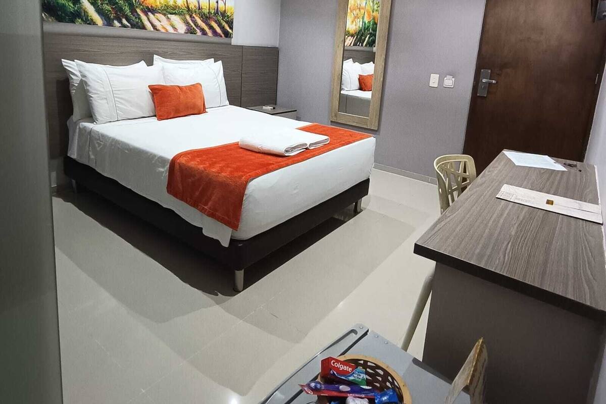 A hotel room with a bed, a desk, a chair, an air conditioning unit, and a painting on the wall.