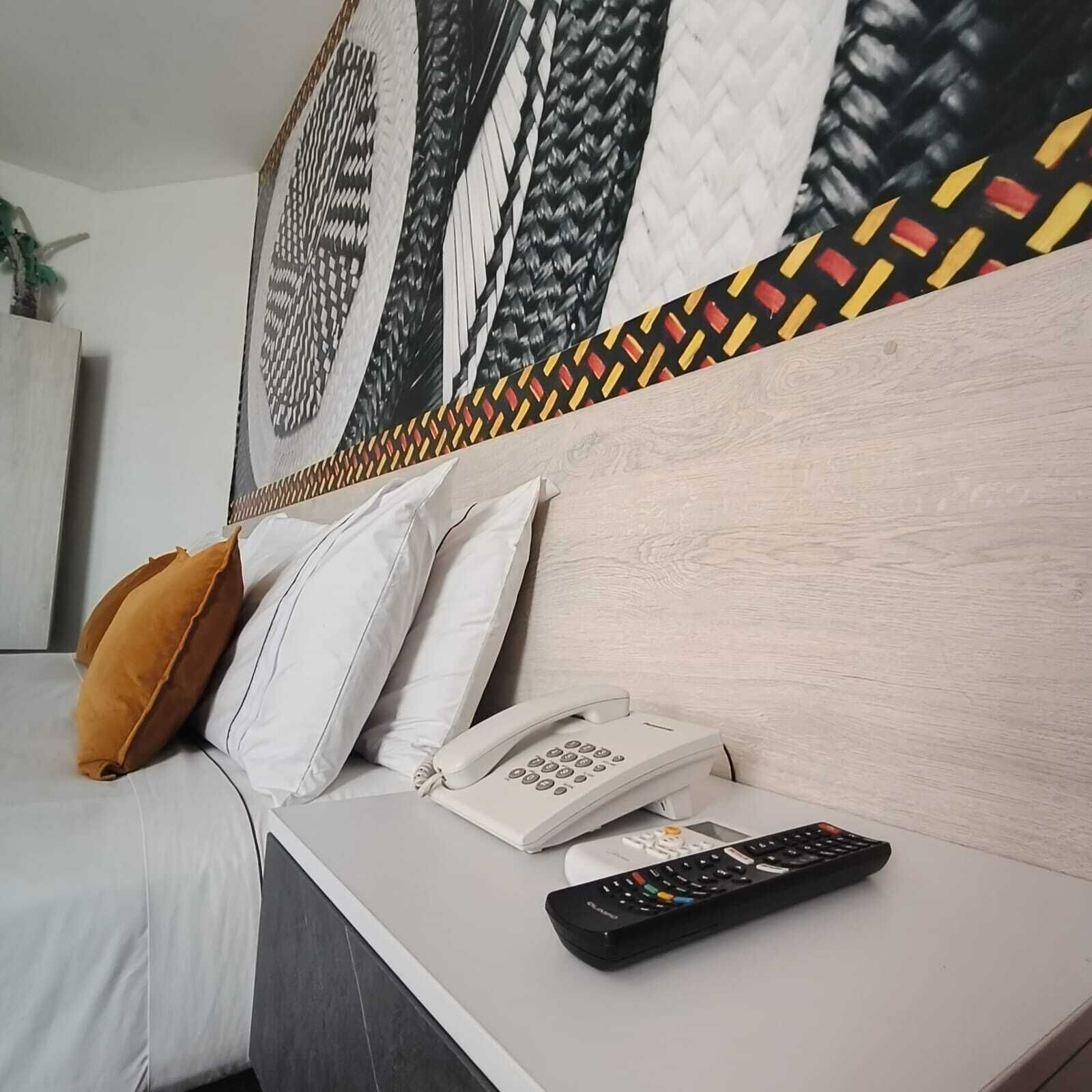 Habitación de hotel con una cama, una mesita de noche con teléfono y control remoto, y una pared con diseño decorativo.