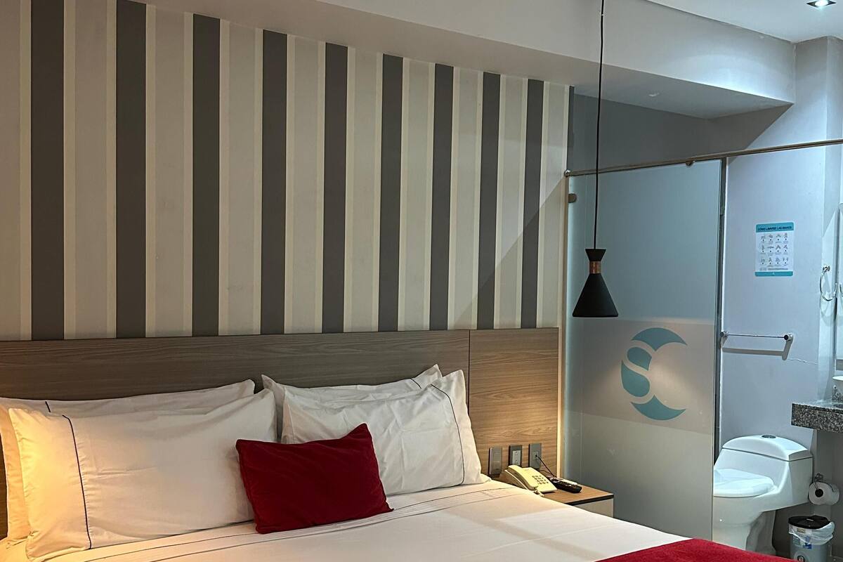 A hotel room with a large bed, a wooden headboard, a red throw blanket, a bedside table with a lamp, and a glass shower enclosure.
