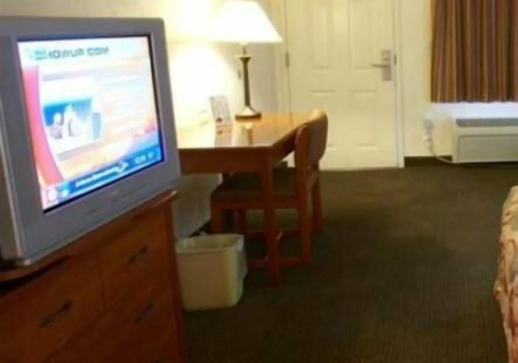 Ein Hotelzimmer mit Fernseher auf einem Holzkasten, einem Schreibtisch mit Stuhl, einer Lampe und einem Bett.