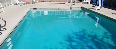 Una piscina rectangular con un cartel de prohibido bucear, rodeada por una valla y sillas de jardín blancas.