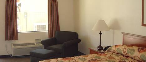 A hotel room with a bed, a chair, an air conditioning unit, a lamp, and a window with curtains.