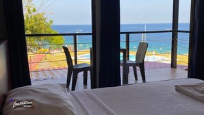 A hotel room with a large window overlooking a pool and the ocean. There is a bed with a white comforter and a folded towel on it. Two chairs are placed near the window. - 24/7 Lantawan Resort (Oslob)