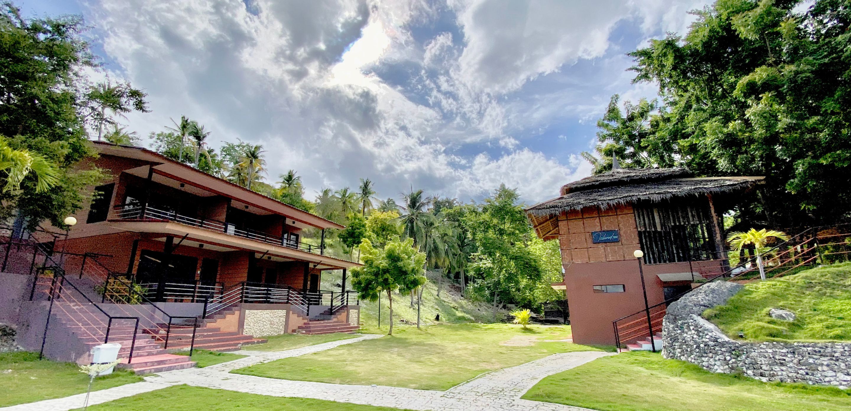 A resort with a two-story building, a thatched-roof structure, and a paved pathway.