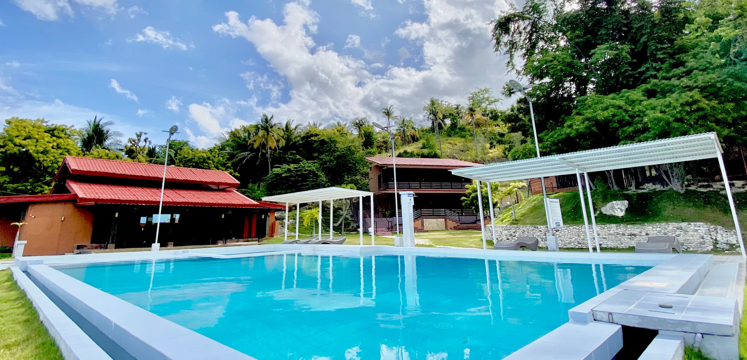 A swimming pool with a white deck, surrounded by a covered seating area and a building with a red roof.