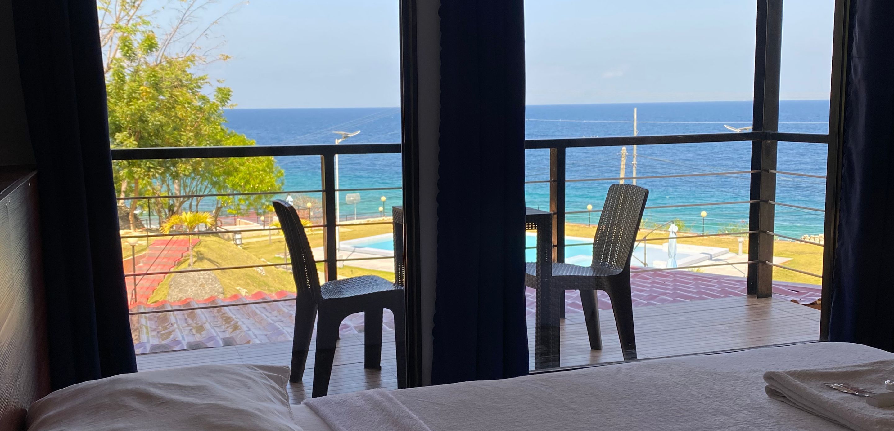 A hotel room with a large window overlooking a pool and the ocean. There is a bed with a white comforter and a folded towel on it. Two chairs are placed near the window.