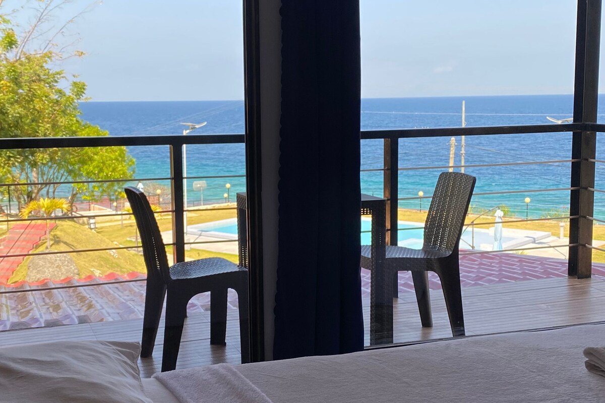 A hotel room with a large window overlooking a pool and the ocean. There is a bed with a white comforter and a folded towel on it. Two chairs are placed near the window.