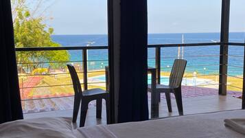 A hotel room with a large window overlooking a pool and the ocean. There is a bed with a white comforter and a folded towel on it. Two chairs are placed near the window.